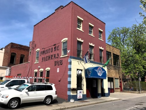 A red brick building with "Founding Fathers Pub" painted on the side, featuring flags and parked vehicles outside.