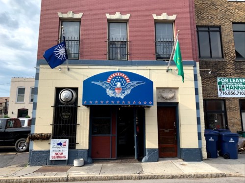 Exterior of a building with a blue awning featuring an eagle emblem, flags, and a sign indicating it's now serving.