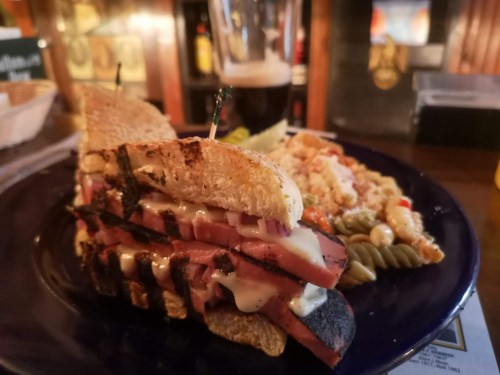 A grilled sandwich with layers of meat and cheese, served with a side of pasta salad and a glass of dark beer.