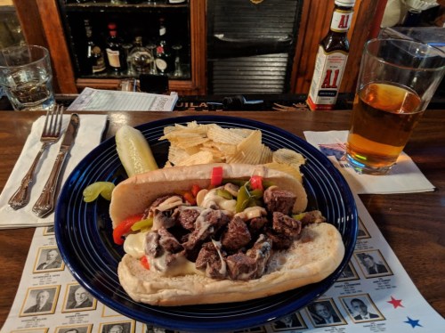 A plate with a sandwich filled with beef, peppers, and onions, served with chips, a pickle, and a glass of beer.