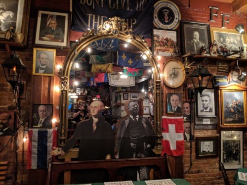 A cozy bar interior filled with historical portraits, flags, and a large mirror reflecting the vibrant decor.