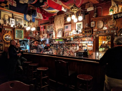 A cozy bar with a wooden counter, vintage decor, and patrons enjoying drinks under hanging lights and flags.