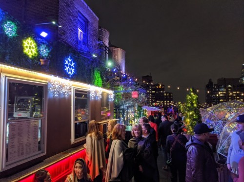 A festive rooftop scene at night, decorated with colorful lights and snowflake ornaments, with a crowd enjoying the atmosphere.