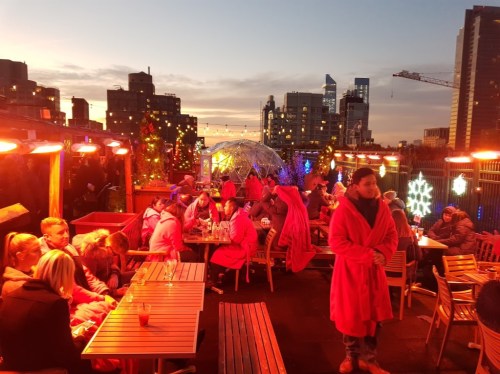 A rooftop bar at sunset, with people in cozy robes enjoying drinks and festive decorations.
