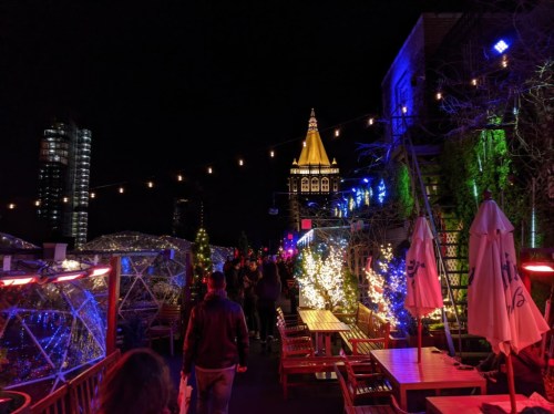 A vibrant rooftop scene at night, adorned with colorful lights and festive decorations, with people walking among tables.