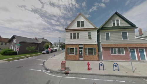 A street view of a two-story building with a storefront, surrounded by residential homes and a fire hydrant nearby.