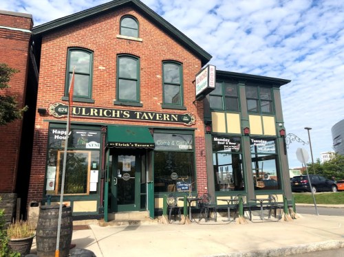 Exterior of Ulrich's Tavern, a brick building with green accents, featuring outdoor seating and a clear blue sky.