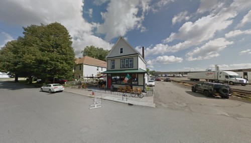 A corner building with outdoor seating, surrounded by trees and parked cars, under a partly cloudy sky.