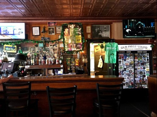 A cozy bar with a wooden counter, shelves of liquor, St. Patrick's Day decorations, and TVs showing sports.