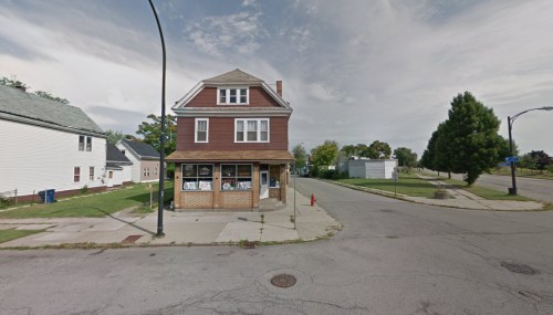A two-story brown building on a street corner, with a storefront and residential homes in the background.
