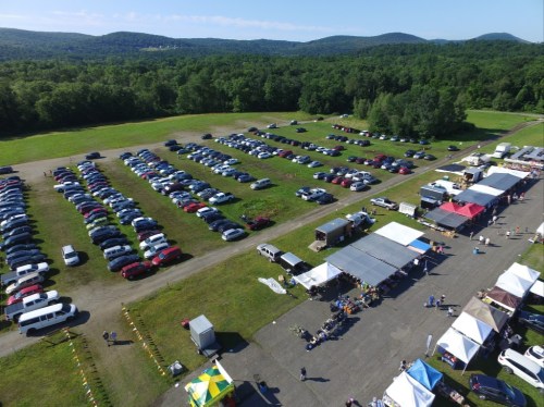 Aerial view of a large parking lot filled with cars, with vendor tents and green hills in the background.