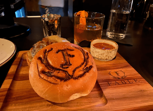 A wooden board with a burger bun featuring a logo, accompanied by a shot glass, a drink, and a small bowl of sauce.