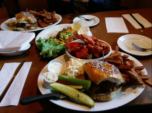 A table with two plates of food: a sandwich with fries, coleslaw, celery, and a platter of wings and vegetables.