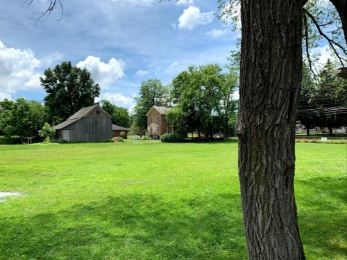 A grassy field with a large tree, a house, and a barn under a blue sky with fluffy clouds.
