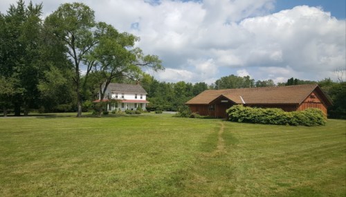A grassy area featuring a white house and a wooden barn under a partly cloudy sky.