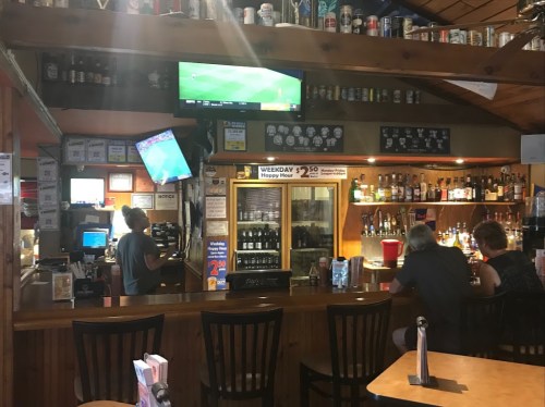 A cozy bar scene with patrons at the counter, a TV showing a sports game, and shelves filled with drinks.
