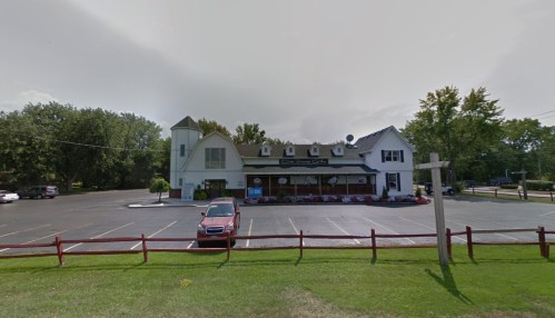 A quaint restaurant with a large parking lot, surrounded by trees and flowers, featuring a sign and a cross in the foreground.