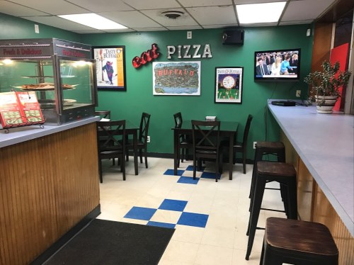 A small pizza shop with green walls, tables, and a TV showing a program. Pizza display case visible on the left.