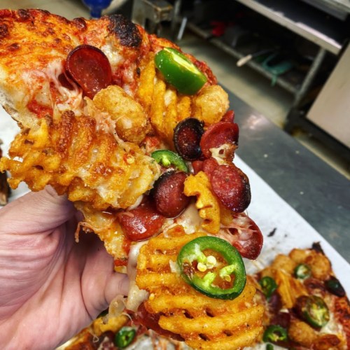 A hand holding a slice of pizza topped with pepperoni, jalapeños, and waffle fries, with melted cheese visible.