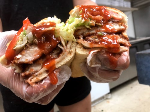 A person holding a sandwich filled with sliced meat, lettuce, and ketchup, wearing gloves in a kitchen setting.