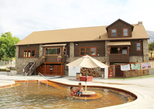 A rustic house with a stone facade and a pool, where two children play near a shaded area with an umbrella.