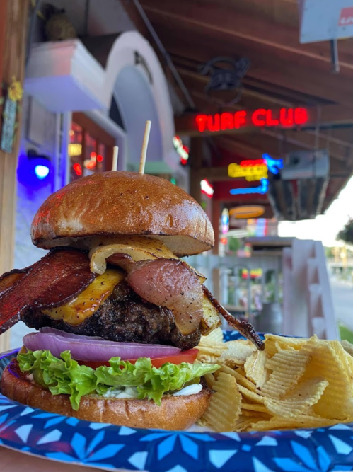 A towering burger with bacon, cheese, and toppings, served with chips, in front of a neon-lit restaurant.