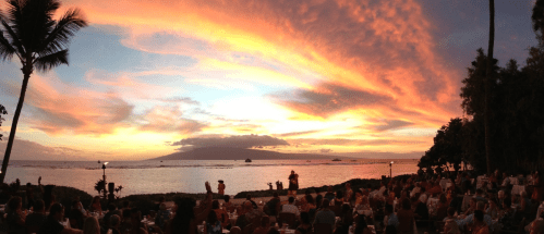 A vibrant sunset over the ocean, with people dining outdoors and palm trees framing the scene.