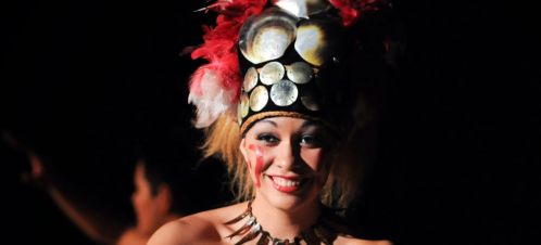 A performer wearing a vibrant headdress with feathers and metallic elements, smiling against a dark background.