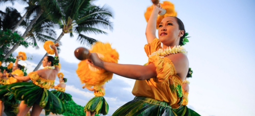 A dancer in a yellow top and green skirt performs hula, holding a wooden instrument, with palm trees in the background.