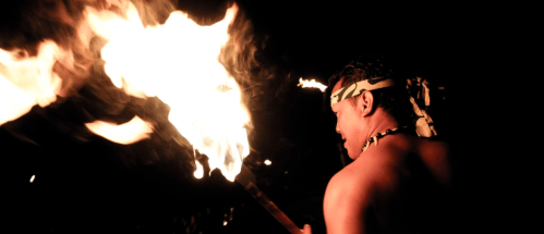 A person with a headband holds a flaming torch, illuminated against a dark background.