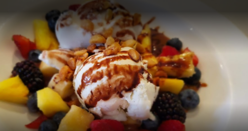 A dessert bowl with two scoops of ice cream topped with chocolate sauce, nuts, and a variety of fresh fruits.