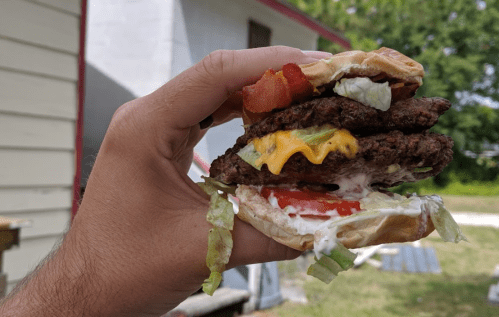 A hand holding a stacked burger with two patties, cheese, bacon, lettuce, tomato, and mayonnaise against a blurred background.