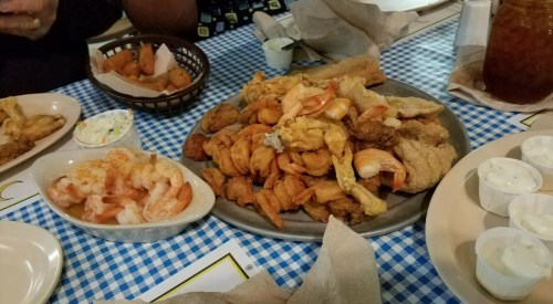 A large platter of fried seafood and chicken, with sides of coleslaw and dipping sauces on a checkered tablecloth.
