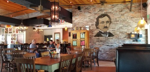 A cozy restaurant interior featuring wooden tables, chairs, and a mural of Edgar Allan Poe on a brick wall.