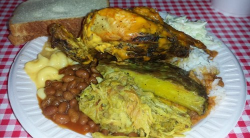 A plate of food featuring grilled chicken, rice, beans, macaroni, collard greens, and a slice of bread on a checkered tablecloth.