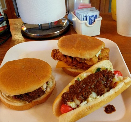 A plate with three sandwiches: a burger, a fried chicken sandwich, and a chili dog with toppings.