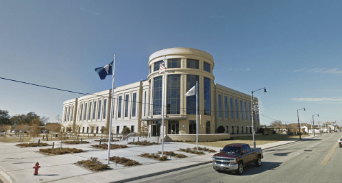 A modern government building with large windows, flags, and a landscaped front, viewed from the street.