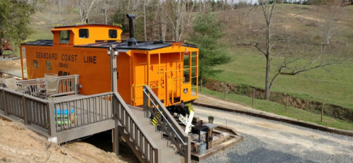 An orange caboose from the Seaboard Coast Line, set on a hillside with a wooden deck and gravel path nearby.