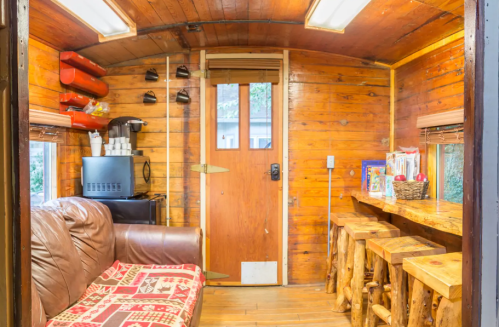 Cozy interior of a wooden cabin with a sofa, bar stools, and a coffee station near the door. Warm, rustic decor.