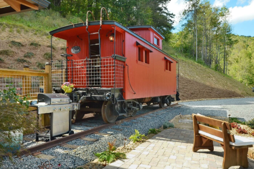 A red caboose on tracks beside a patio area with a grill and seating, surrounded by greenery and a hillside.