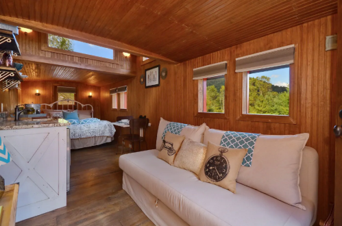 Cozy interior of a wooden cabin featuring a sofa, decorative pillows, and a view of a bedroom area with windows.
