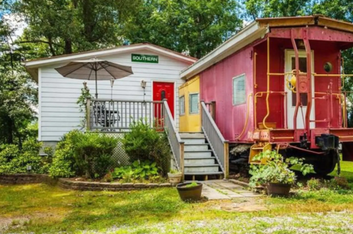 A colorful train caboose attached to a white house with a red door and an umbrella on the porch, surrounded by greenery.