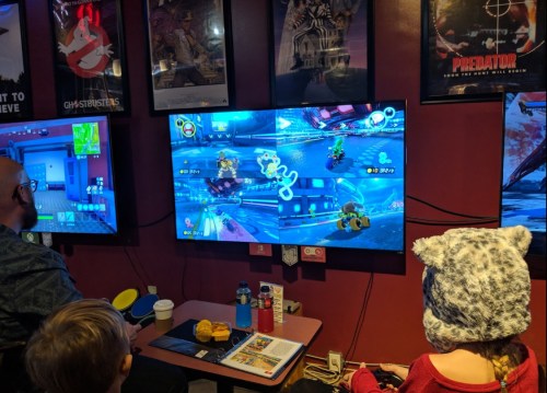 Two children and an adult play a racing video game on a large screen, surrounded by movie posters.