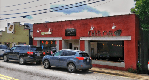 A red brick building with "GAZE" and "Odd's Cafe" signs, parked cars in front, and a cloudy sky above.