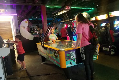 A girl plays air hockey while a boy interacts with an arcade machine in a vibrant game room filled with colorful lights.
