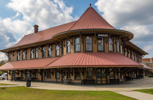 Historic train station with a distinctive red roof and large windows, set against a cloudy sky in Hamlet, NC.
