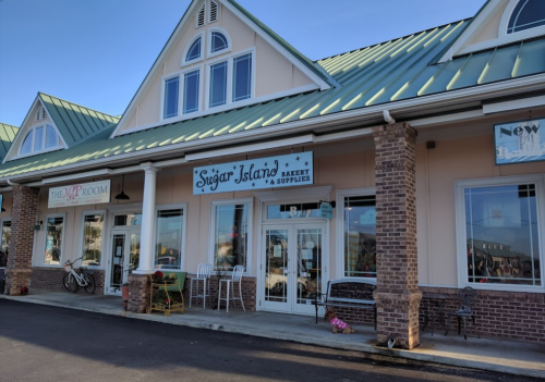 Exterior of a bakery named "Sugar Island" with a green roof and large windows, surrounded by other shops.