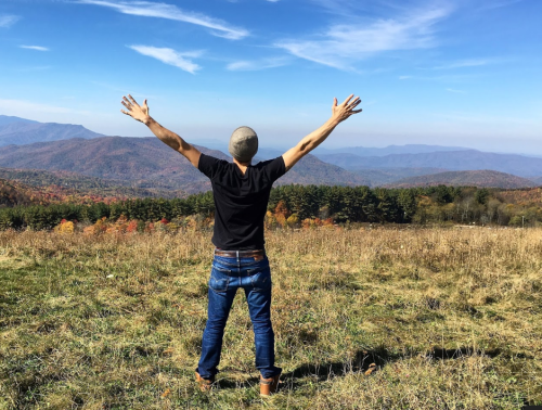 A person stands with arms outstretched, overlooking a scenic mountain landscape with colorful autumn foliage.