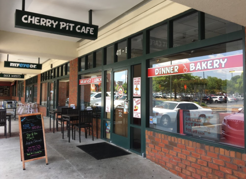 Exterior view of Cherry Pit Cafe with outdoor seating, featuring signs for breakfast, lunch, dinner, and bakery items.