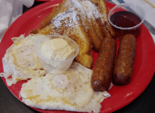 A red plate with two fried eggs, French toast, sausage links, and a small cup of syrup.